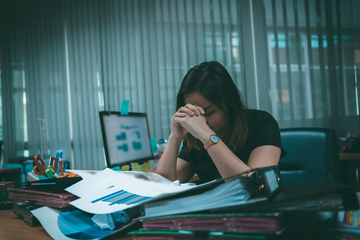 IT issues causing employee stress and productivity loss as a frustrated woman sits at a cluttered desk with her head in her hands in front of a malfunctioning computer.